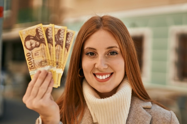 Foto gratuita joven pelirroja sonriendo feliz sosteniendo billetes de florín húngaro en la ciudad
