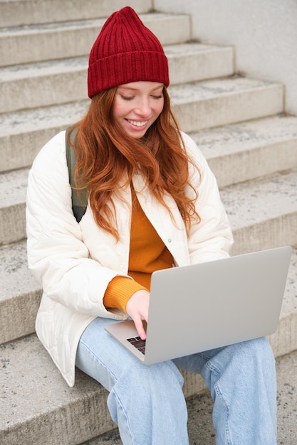 Una joven pelirroja sonriente escribiendo en el teclado de una computadora portátil sentada al aire libre en las escaleras con una computadora