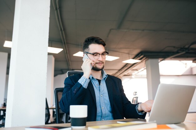 Joven de pelo oscuro está trabajando en la mesa en la oficina. Viste camisa azul con chaqueta negra. Él está hablando por teléfono y mostrando a la computadora portátil.