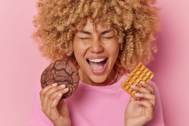 Una joven de pelo rizado muy contenta sostiene una deliciosa galleta de chocolate y un gofre casero tiene un diente dulce tiene adicción al azúcar exclama en voz alta mantiene la boca abierta aislada sobre un fondo rosado
