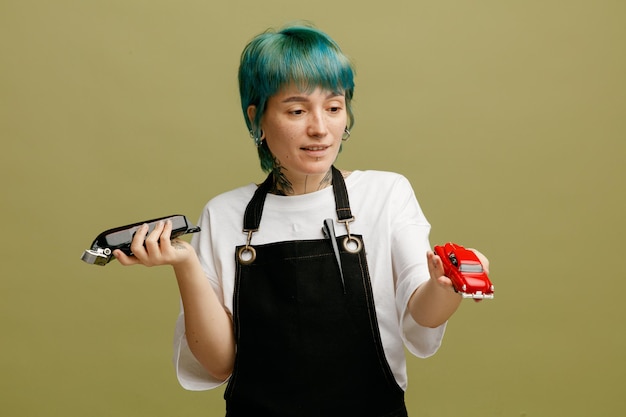 Una joven peluquera complacida con uniforme sosteniendo una cortadora de cabello y una figurilla de un auto mirando una figurita de un auto aislada en un fondo verde oliva