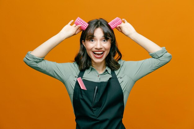 Joven peluquera con delantal con peine sosteniendo rulos sobre su cabeza como orejas de conejo mirando a la cámara feliz y positiva sonriendo alegremente de pie sobre fondo naranja