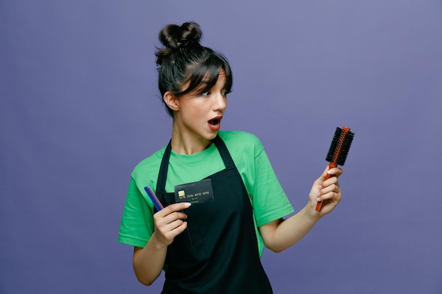 Joven peluquera con delantal sosteniendo un cepillo para el cabello y una tarjeta de crédito que parece confundida y sorprendida de pie sobre un fondo azul