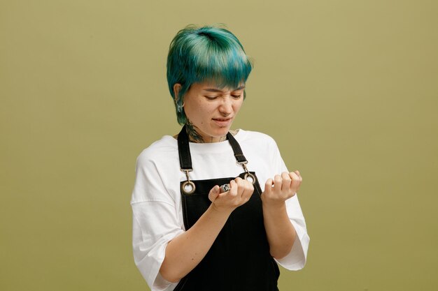 Una joven peluquera disgustada con uniforme sosteniendo cortauñas mirando sus uñas aisladas en un fondo verde oliva