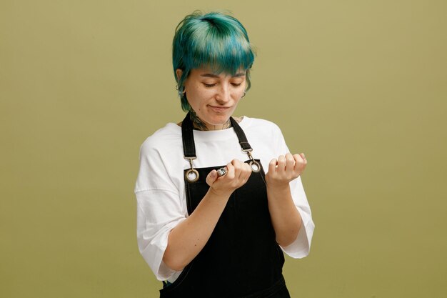 Una joven peluquera disgustada con uniforme sosteniendo cortauñas mirando sus uñas aisladas en un fondo verde oliva