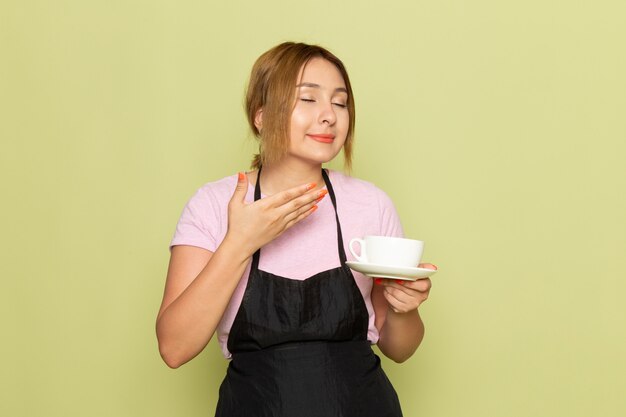 Una joven peluquera de vista frontal en camiseta rosa y capa negra sosteniendo la taza sonriendo en verde