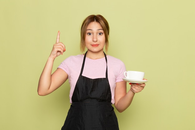 Una joven peluquera de vista frontal en camiseta rosa y capa negra sosteniendo la taza sonriendo en verde