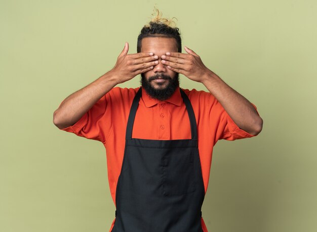 Joven peluquero afroamericano vistiendo uniforme cubriendo los ojos con las manos