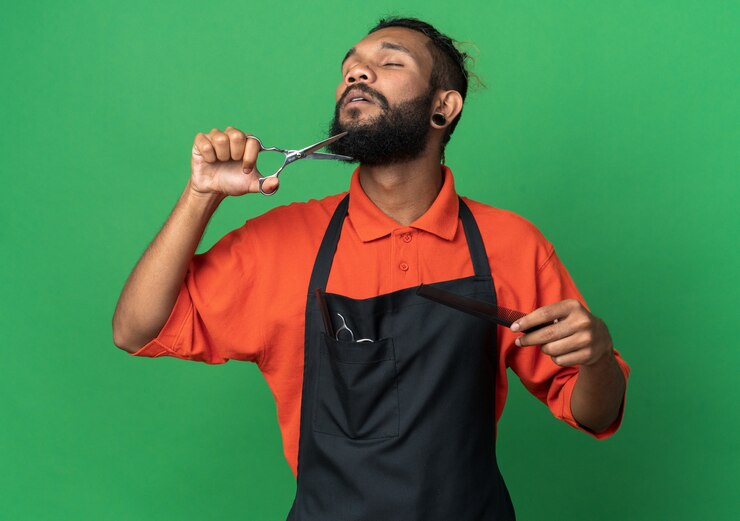 Foto gratuito joven peluquero afroamericano vistiendo uniforme sosteniendo peine y tijeras cortando su barba con los ojos cerrados aislado en la pared verde