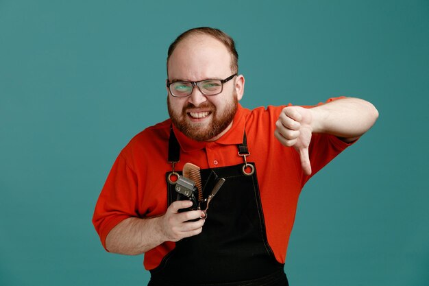 Joven peluquero descontento con gafas, camisa roja y delantal de peluquero sosteniendo herramientas de barbería mirando a la cámara mostrando el pulgar hacia abajo aislado en el fondo azul