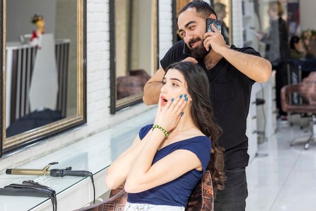 Joven peluquero hablando por teléfono y su cliente está en estado de shock Foto de alta calidad