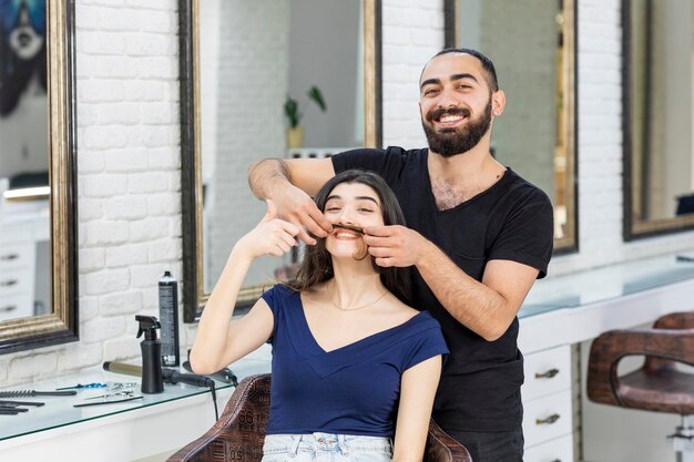 Joven peluquero sosteniendo el cabello de una mujer como bigote y ambos riéndose Foto de alta calidad