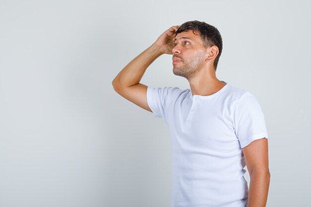 Joven pensando con la mano en la cabeza en camiseta blanca, vista frontal.