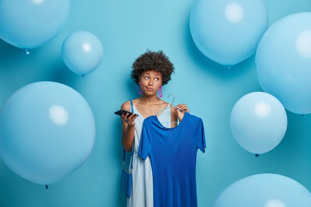 Una joven pensativa de piel oscura tiene el pelo rizado, sostiene un elegante vestido azul en una percha, un teléfono móvil en la mano, se viste para una fiesta temática azul, mira a un lado, posa contra globos con una mirada pensativa