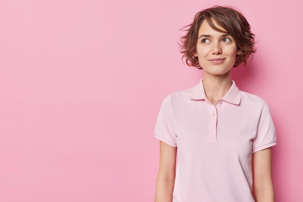 Una joven pensativa y soñadora con un peinado corto concentrado tiene una expresión soñadora que considera que algo lleva una camiseta informal aislada sobre un fondo rosa. Concepto de personas y pensamientos.