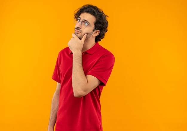 Foto gratuita joven pensativo en camisa roja con gafas ópticas pone la mano en la barbilla aislada en la pared naranja