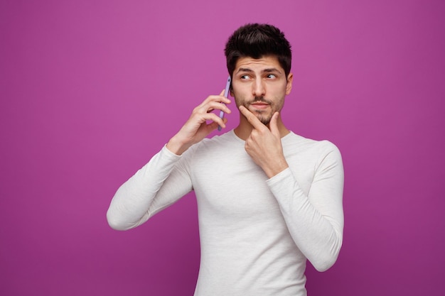 Un joven pensativo con la mano en la barbilla mirando a un lado mientras habla por teléfono aislado en un fondo morado
