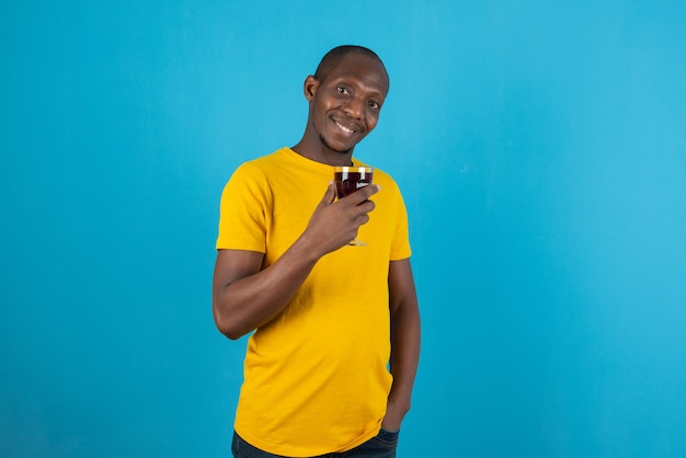 Joven de piel oscura con camisa amarilla sosteniendo una copa de vino en la pared azul