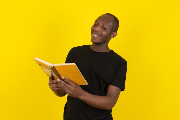 Joven de piel oscura leyendo un libro interesante en la pared amarilla
