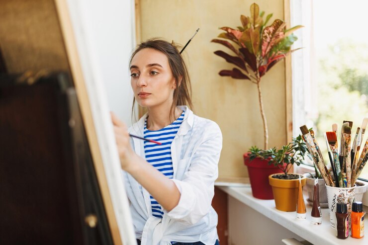 Foto gratuito joven pintor pensativo con camisa blanca y camiseta a rayas dibujando soñadoramente en caballete cerca de la ventana en casa