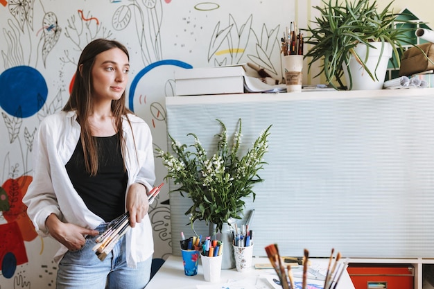 Foto gratuita joven pintora bonita sosteniendo pinceles en la mano mientras mira soñadoramente a un lado con grandes patrones de lienzo en el fondo de casa