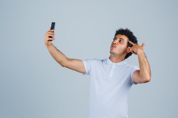 Joven posando mientras toma selfie en teléfono móvil con camiseta blanca y mirando confiado, vista frontal.