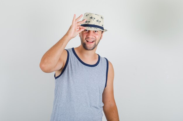 Joven posando sosteniendo su sombrero en camiseta gris y luciendo alegre.