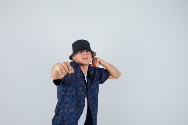 Joven en pose de lucha en camiseta blanca, camisa floral, gorra y mirando confiado. vista frontal.