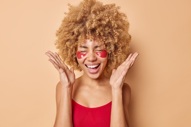 Una joven positiva con el pelo rizado mantiene las palmas levantadas, se siente encantada, se ríe alegremente, tiene un estado de ánimo optimista y aplica parches de hidrogel debajo de los ojos para reducir la hinchazón aislada sobre un fondo beige.