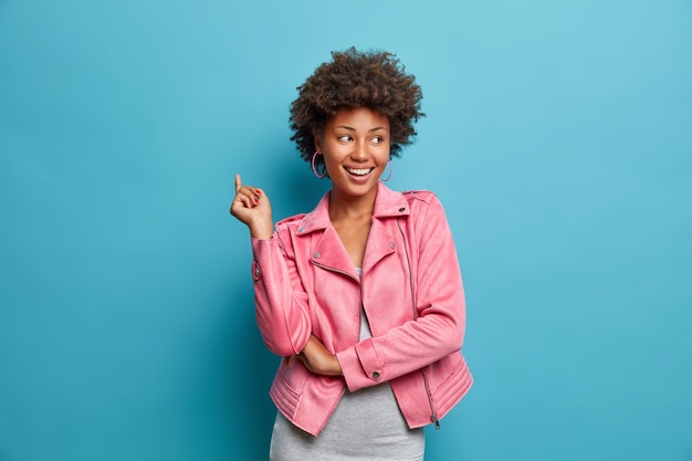 Foto gratuita la joven positiva de piel oscura se ríe positivamente, tiene una expresión de rostro alegre y emocionada, levanta la mano y se entretiene con amigos, usa una chaqueta elegante,