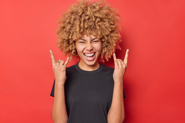 Una joven positiva y rizada muestra un gesto de cuerno que disfruta de la música rock y viste una camiseta negra casual aislada sobre un fondo rojo vívido