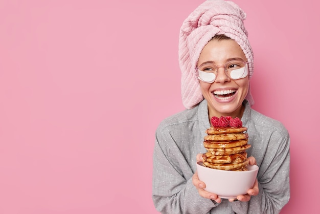 Joven positiva sostiene un tazón con un montón de deliciosos panqueques, sonríe con dientes concentrados, aplica parches de belleza para reducir las arrugas, se siente refrescado después de dormir, usa pijamas en el interior