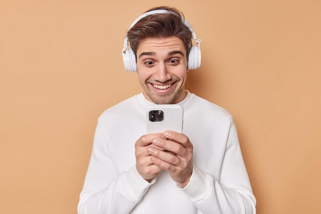 El joven positivo mira felizmente la pantalla del teléfono inteligente, lee una historia divertida, deja comentarios debajo de la publicación, escucha la pista de audio a través de auriculares inalámbricos, vestido con un jersey casual aislado sobre fondo beige