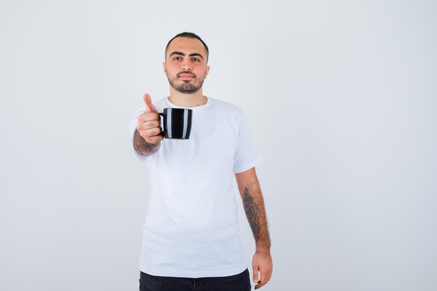 Joven presentando taza de té en camiseta blanca y pantalón negro y mirando feliz