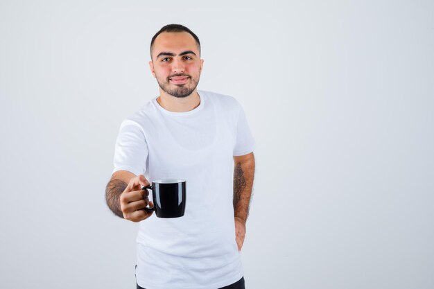 Joven presentando taza de té en camiseta blanca y pantalón negro y mirando feliz