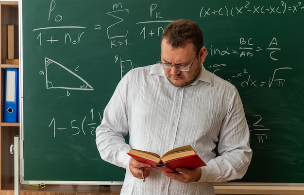 Joven profesor concentrado con gafas de pie delante de la pizarra en el aula sosteniendo un libro de lectura de palo puntero