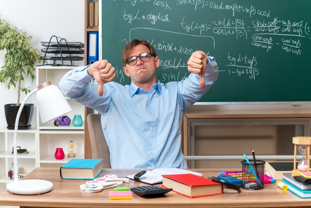 Joven profesor con gafas disgustado mostrando los pulgares hacia abajo sentado en el escritorio de la escuela con libros y notas frente a la pizarra en el aula