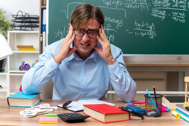 Foto gratuita joven profesor con gafas estresado y nervioso sentado en el escritorio de la escuela con libros y notas delante de la pizarra en el aula