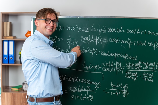 Joven profesor con gafas explicando la lección feliz y positiva sonriendo de pie cerca de la pizarra con fórmulas matemáticas en el aula