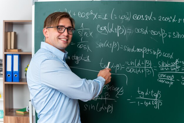 Joven profesor con gafas explicando la lección feliz y positiva sonriendo de pie cerca de la pizarra con fórmulas matemáticas en el aula