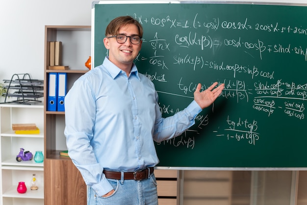 Joven profesor con gafas feliz y positivo explicando la lección de pie cerca de la pizarra con fórmulas matemáticas en el aula