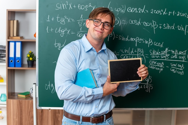 Foto gratuita joven profesor con gafas con libro sosteniendo una pequeña pizarra mirando a la cámara sonriendo confiados de pie cerca de la pizarra con fórmulas matemáticas en el aula