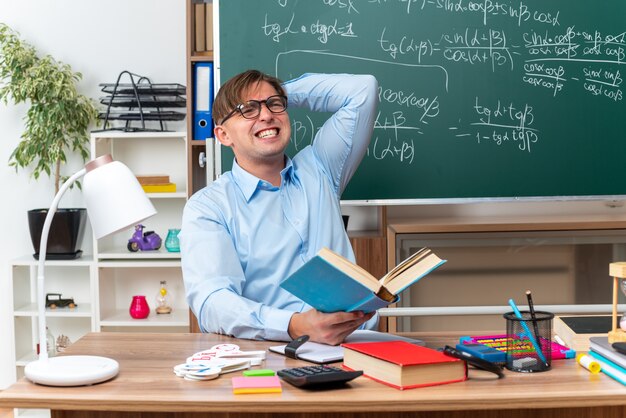 Joven profesor con gafas mirando confundido y decepcionado sentado en el escritorio de la escuela con libros y notas frente a la pizarra en el aula