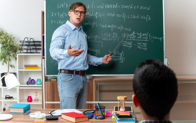 Joven profesor con gafas mirando confundido y disgustado explicando la lección a los alumnos, de pie cerca de la pizarra con fórmulas matemáticas en el aula