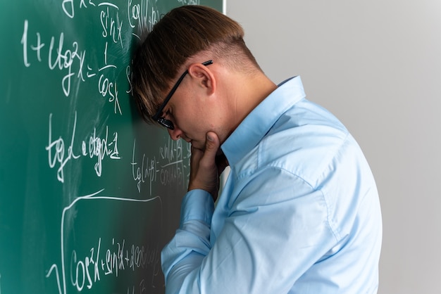 Joven profesor con gafas mirando desconcertado al no tener respuesta de pie cerca de la pizarra con fórmulas matemáticas en el aula