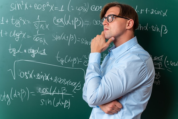 Joven profesor con gafas mirando a un lado con la mano cerca de la barbilla con expresión pensativa pensando de pie cerca de la pizarra con fórmulas matemáticas en el aula