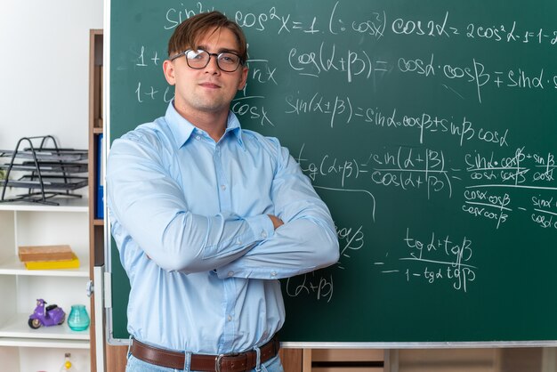 Joven profesor con gafas mirando con sonrisa de confianza en la cara con los brazos cruzados de pie cerca de la pizarra con fórmulas matemáticas en el aula