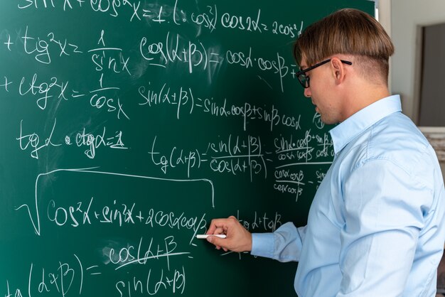 Joven profesor con gafas de pie cerca de la pizarra escribiendo en fórmulas matemáticas de pizarra explicando la lección que parece seguro en el aula