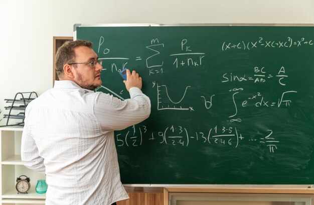 Joven profesor con gafas de pie detrás de la vista delante de la pizarra en el aula mirando al lado pizarra de limpieza con borrador de tiza