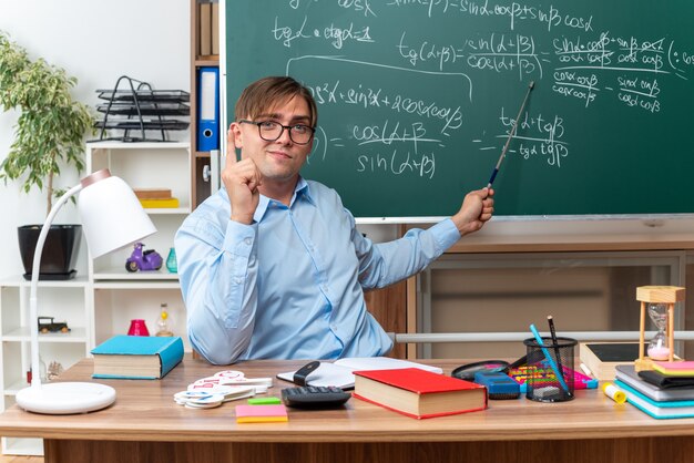 Joven profesor con gafas con puntero explicando la lección mirando confiado sentado en el escritorio de la escuela con libros y notas frente a la pizarra en el aula